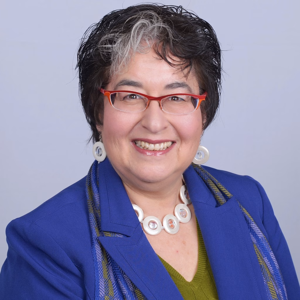 Betty, a person with short, curly, salt-and-pepper hair. She is smiling and wearing red glasses, a blue blazer, a green top, and white circular jewelry. Behind her is a plain, light-colored background.