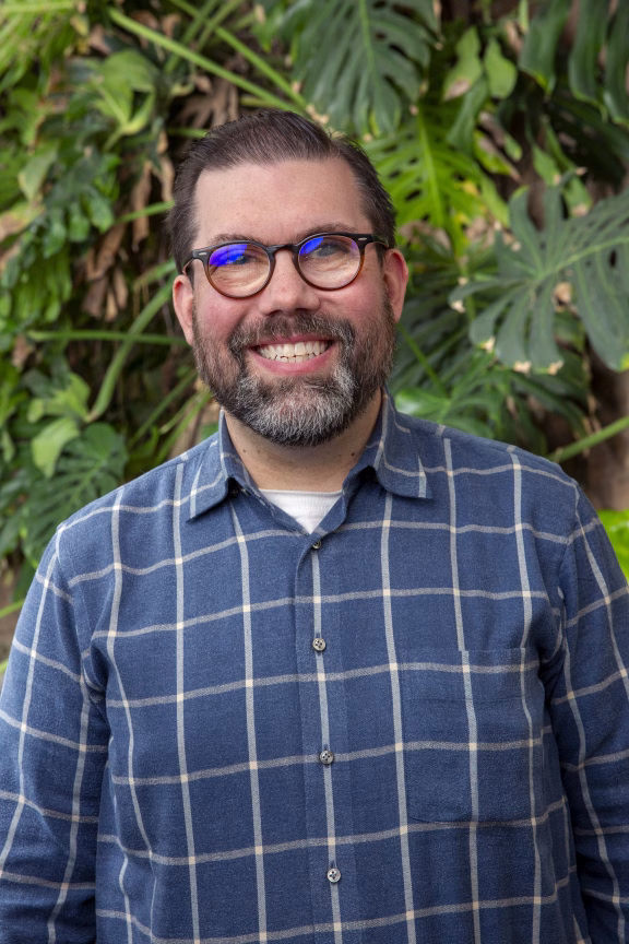 Bill, a smiling person with glasses and a beard. He is wearing a blue plaid shirt and stands in front of green leafy plants.