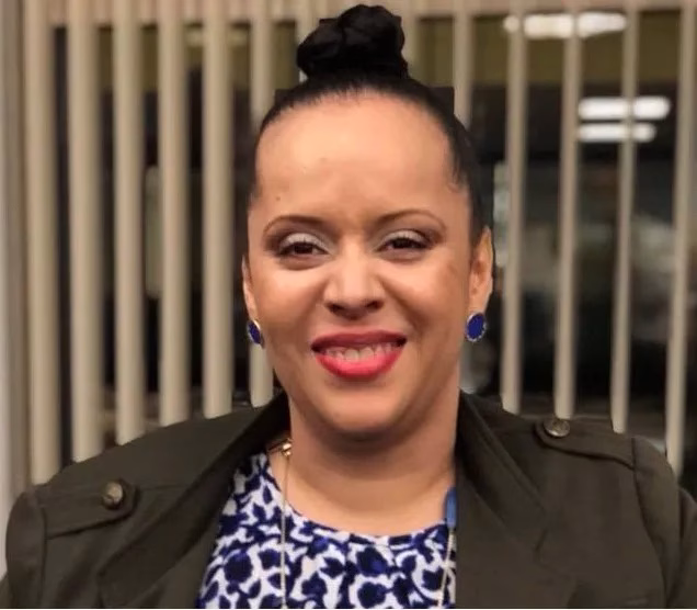Heather is a light-complected Black woman shown shoulders up wearing hair in bun atop her head, blue button earrings, makeup with red lipstick and smiling. She is wearing olive-colored blazer and blue and white patterned blouse with long necklace of various blue-colored pendants