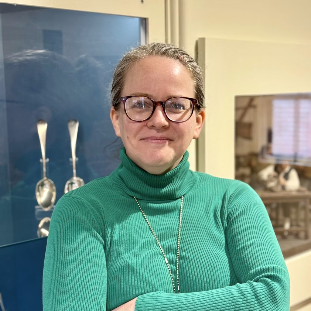 Kristin, a person wearing glasses and a green turtleneck sweater. She is smiling with her arms crossed, standing in front of a display case with silver spoons and a diorama behind her.