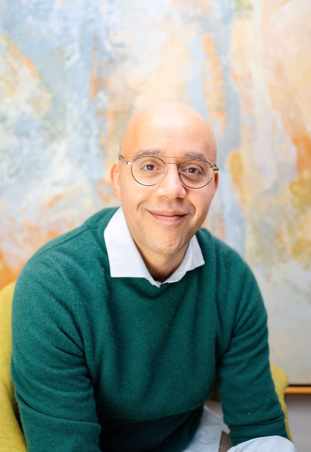 A light-skinned black male wearing a green sweater over a collar shirt with glasses and sitting in front of a pastel artwork.