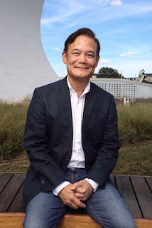 Roger, a person wearing a dark blazer and jeans, sits on a bench outdoors, smiling at the camera. Behind him are tall grasses and a curved white structure beneath a blue sky.