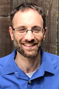 Ross, a person with short brown hair, a beard, and glasses. He is smiling and wearing a blue collared shirt. Behind him is a wooden background.