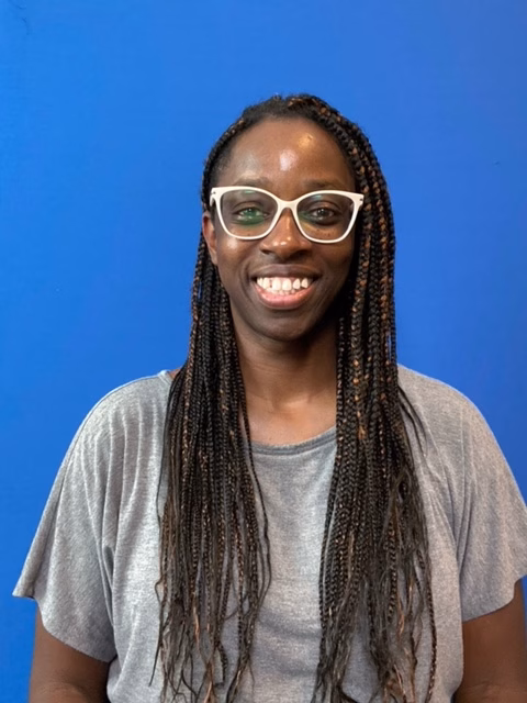 Stephanie, a person with long braids and white-framed glasses. She is smiling and wearing a loose gray t-shirt. Behind her is a solid blue background.