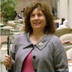 Valerie, a person with curly brown hair, wearing a gray jacket over a pink top. She is smiling and standing indoors in a seating area. She holds a mobility cane.
