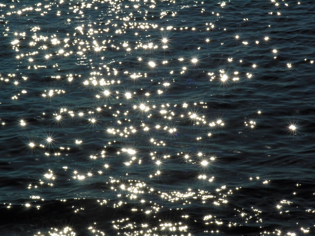 A landscape image entirely composed of deep blue waves that ripple with shades darkening to the right and bottom of the photo. Hundreds of sparkling bursts of light are sprinkled randomly throughout slightly more concentrated on the left. Several of the individual bursts resemble the sun with rays of light extending from their round center points.